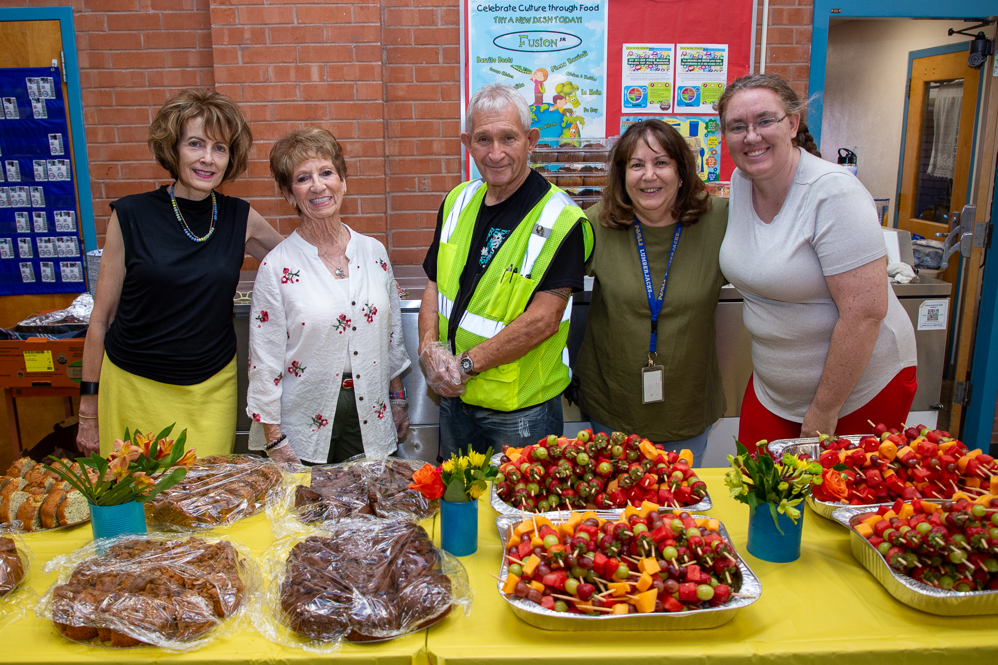 A group of people smile behind a table filled with breakfast pastries and fruit