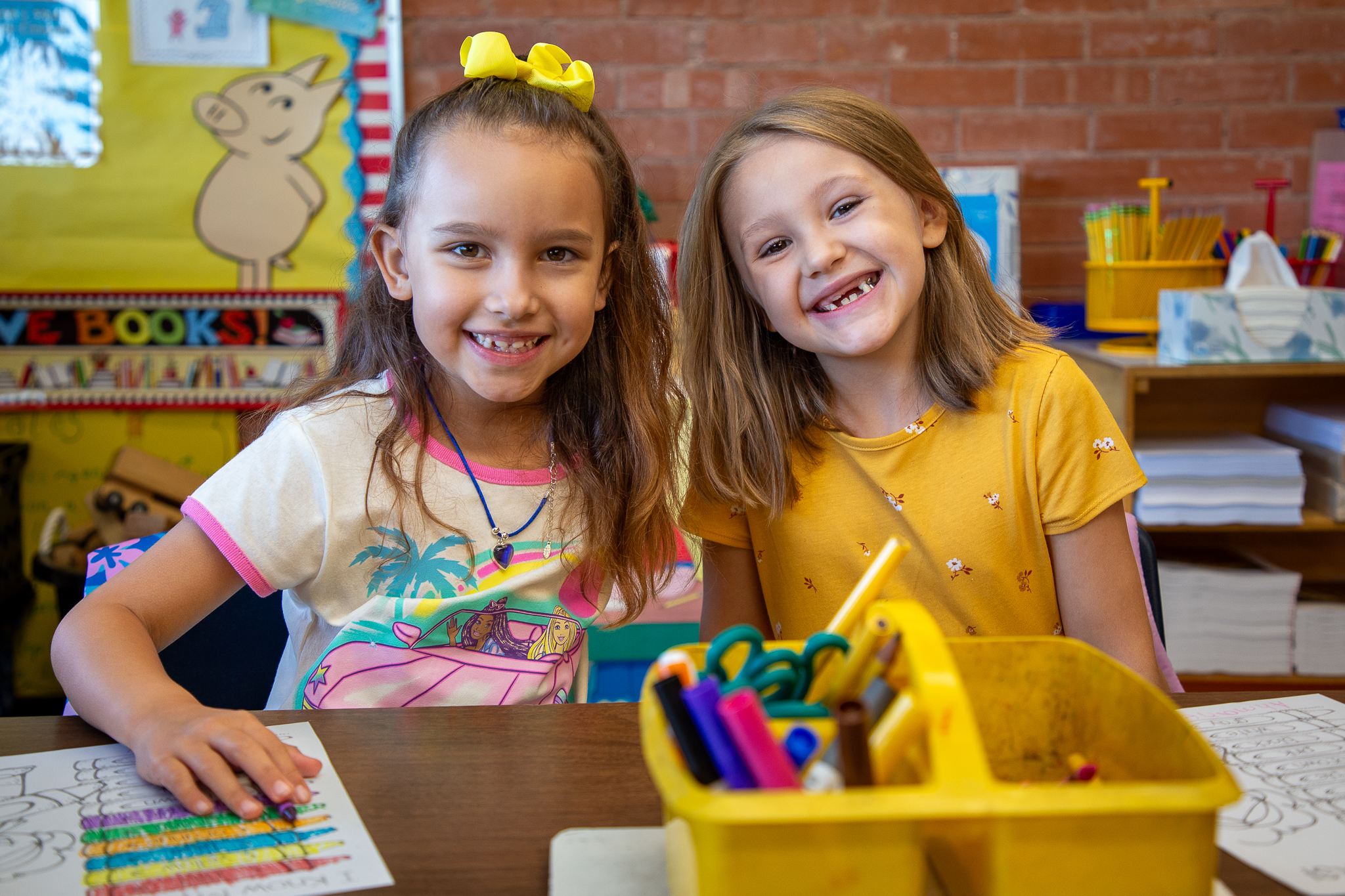 Two girls smile at their desk with school supplies in the foreground