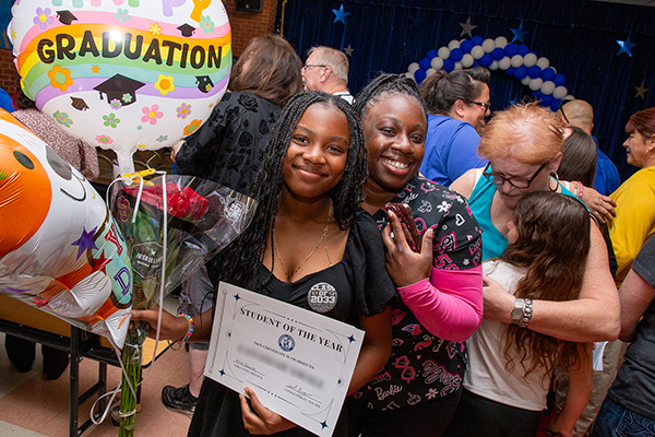 A girl holds a bunch of graduation balloons and smiles next to her mom