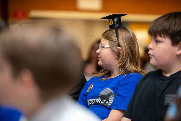 A girl wearing a graduation headband and a boy watch the ceremony