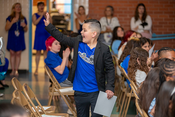 A boy waves to his classmates as he holds his promotion certificate