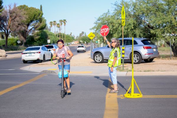 A girl rides her scooter as the crossing guard walks her across the street