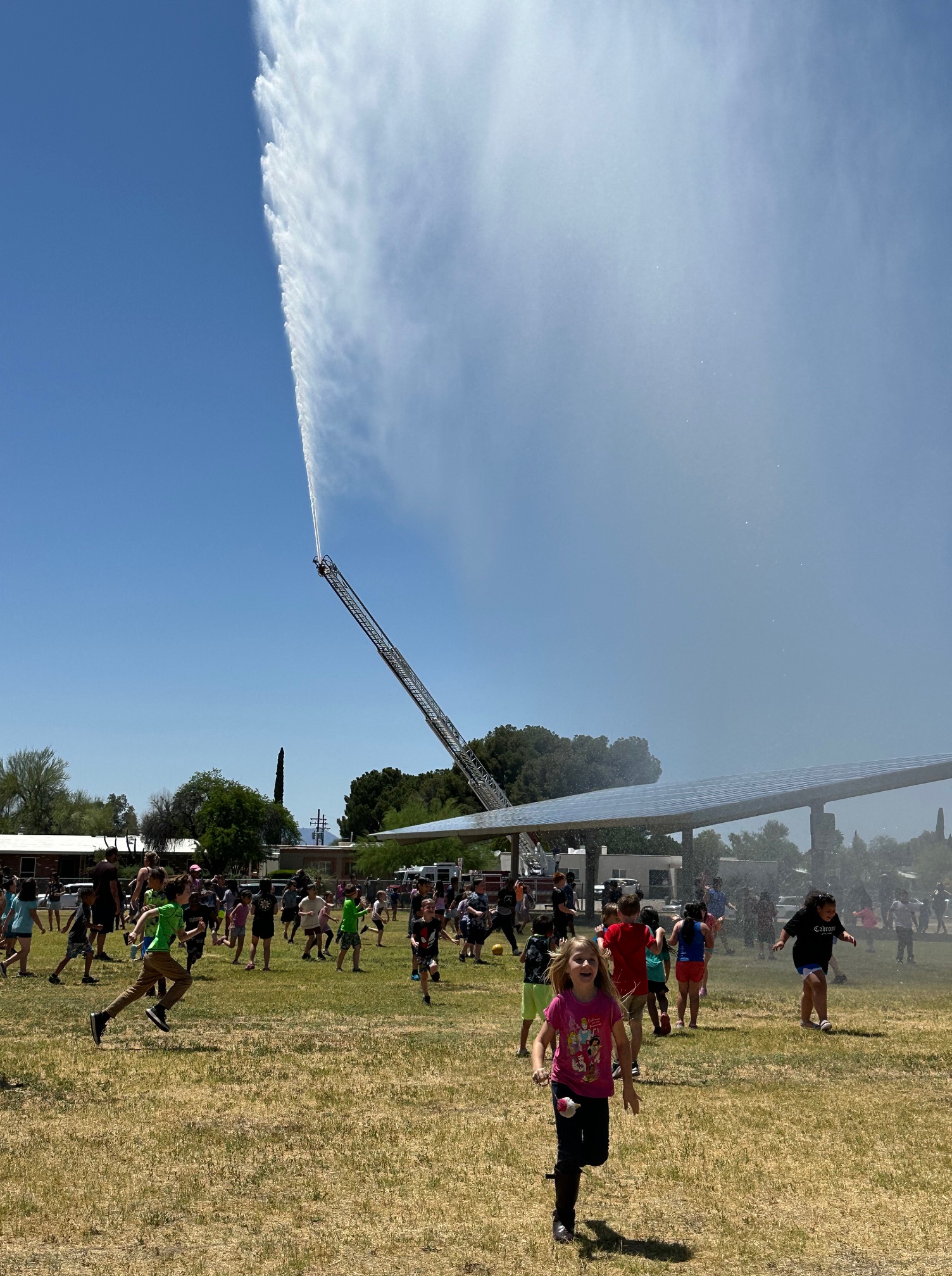 Students run around on the field outside while a fire truck sprays them with water